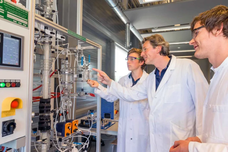Drei Personen in Laborkitteln und mit Schutzbrillen untersuchen und diskutieren eine komplexe Mischanlage mit Rohren, Schläuchen und Schalttafeln in einem modernen Industrielabor. Eine Person zeigt auf das Gerät.