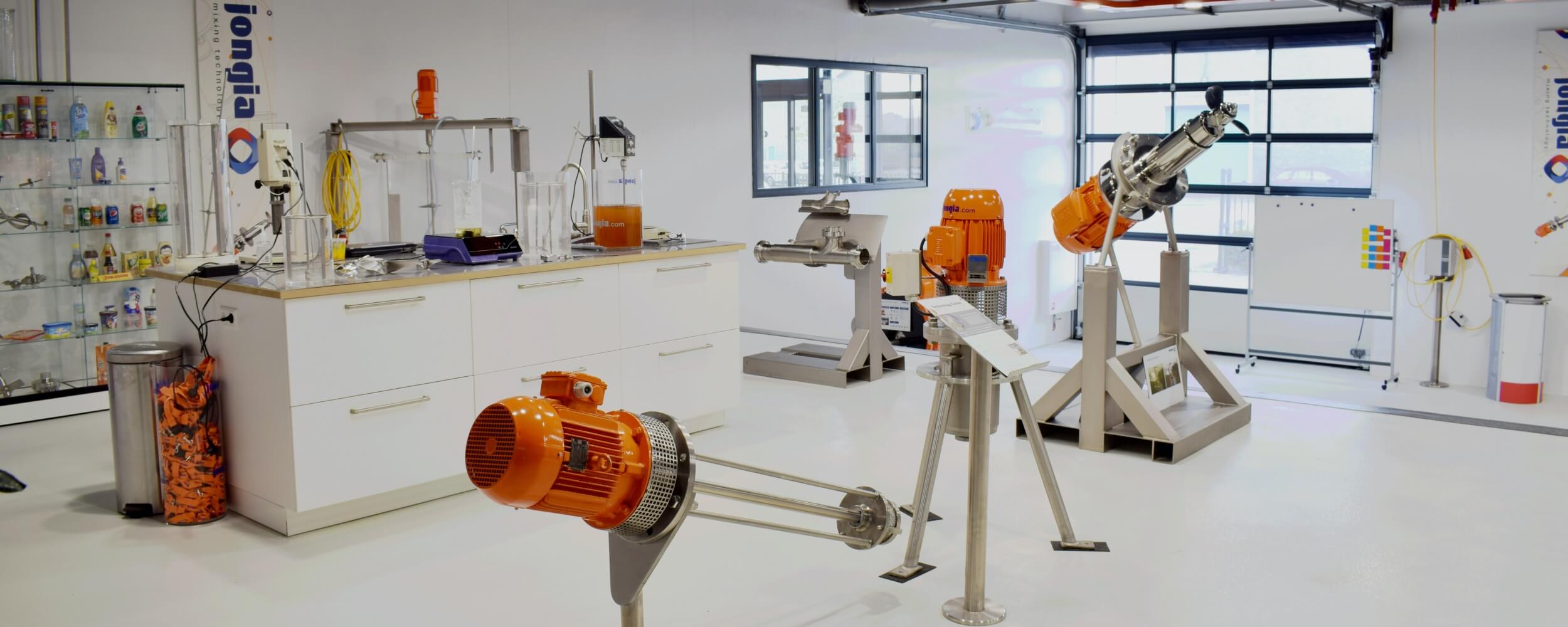 A bright, modern laboratory displaying various orange industrial mixing machines, cabinets with scientific equipment, glass shelves with bottles, and large windows letting in natural light for optimal mixing operations.