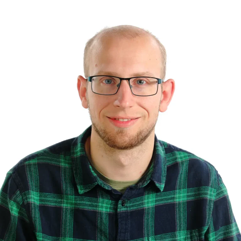 A man with short light hair, glasses, and a trimmed beard smiles at the camera. He is wearing a green and black plaid shirt, mixing bold patterns against a plain white background.