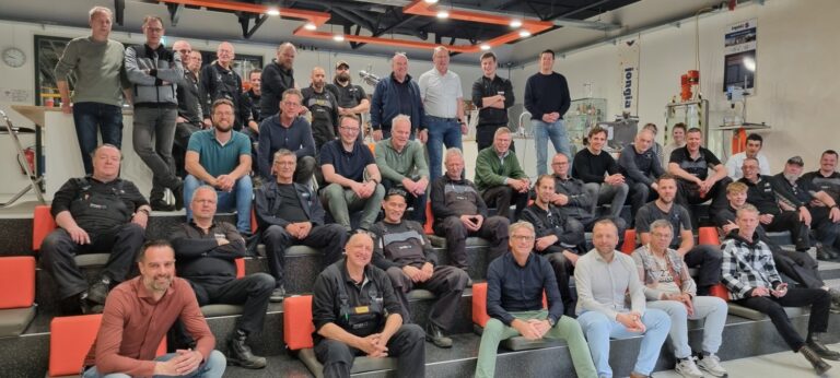 A large group of people, mostly men, sit and stand on tiered steps inside an industrial or workshop setting, mixing camaraderie and teamwork as they pose and smile for a group photo.