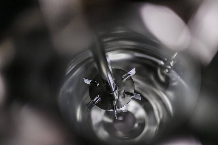 Nahaufnahme eines metallisch glänzenden Industriemischers mit Mischflügeln und einem zentralen Stab in einem zylindrischen Behälter, von oben gesehen mit Spiegelungen und unscharfen Kanten.