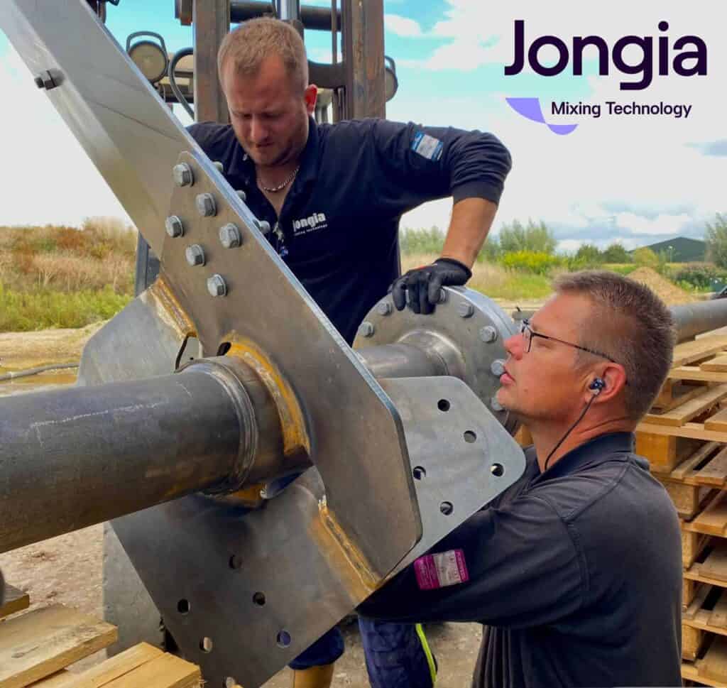 Zwei Männer in schwarzen Jongia-Uniformen bauen draußen große Metallmaschinen zusammen. Der eine stellt die Bolzen ein, während der andere genau beobachtet. Das Logo von Jongia Mixing Technology befindet sich oben rechts. Der Hintergrund zeigt bewölkten Himmel und Grün.