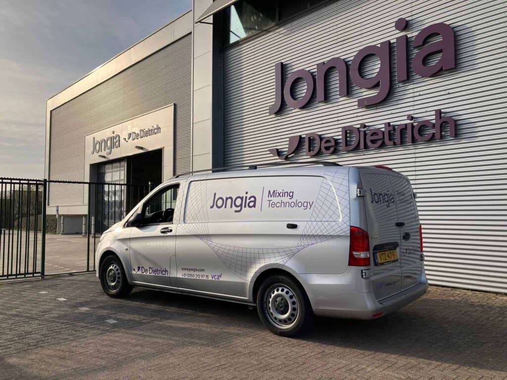 Ein silberner Van mit Jongia Mixing Technology und De Dietrich-Logos steht vor einem großen Industriegebäude mit passender Beschilderung. Der Wagen befindet sich in der Nähe eines Eingangstors und das Hauptgebäude ist mit silbernen Tafeln mit dem Logo von Jongia / De Dietrich verkleidet.