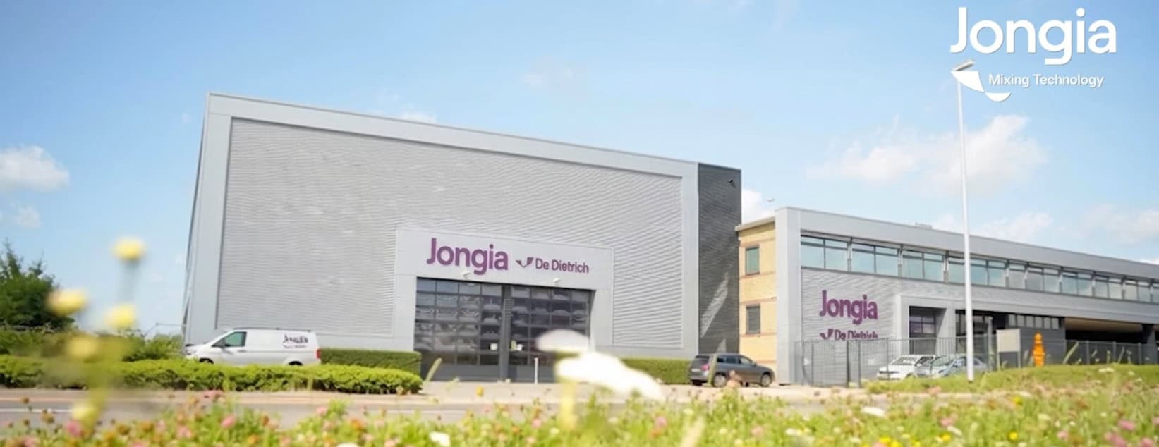 Modern industrial building with Jongia De Dietrich signage on two sections. A white van is parked nearby. Blue sky, grass, and wildflowers are in the foreground. Jongia Mixing Technology logo appears in the top right corner.