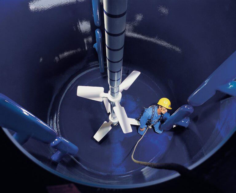 A worker in a yellow hard hat and blue uniform is inside a large, shiny blue industrial tank. A giant white mixing impeller is suspended above them, attached to a vertical shaft. The worker holds a safety rope and looks upward, surrounded by smooth tank walls.