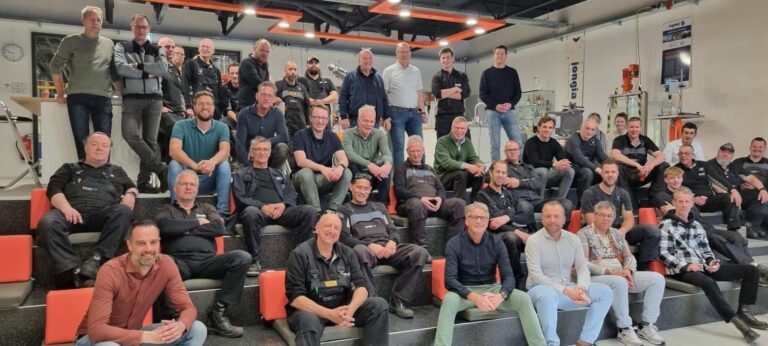 Een grote groep mensen, voornamelijk mannen, zit en staat op traptreden in een moderne, industriële binnenomgeving. Ze zijn casual gekleed, sommigen dragen uniformen. De achtergrond toont technische apparatuur en kantoorelementen. Iedereen kijkt naar de camera en glimlacht.