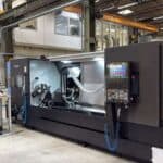 A large CNC machine stands in an industrial workshop with tools and equipment nearby. The machine has a digital control panel, and the Jongia Mixing Technology logo is visible on the wall. Overhead cranes and shelving are in the background.