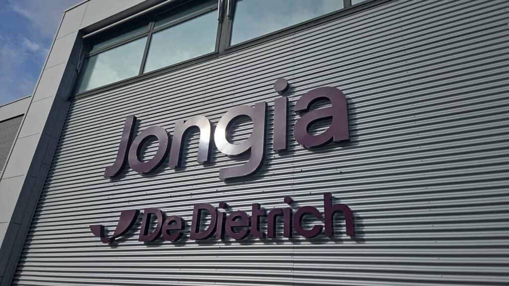 Een gevel van gegolfd metalen gebouw met grote, donkerpaarse 3D-letters die "Jongia" spellen boven een kleiner "De Dietrich"-logo en tekst. Het gebouw heeft grote ramen boven het bord en een blauwe lucht is zichtbaar in de linkerbovenhoek.