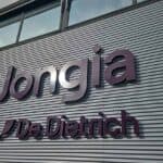 A corrugated metal building facade with large, dark purple 3D letters spelling “Jongia” above a smaller “De Dietrich” logo and text. The building has large windows above the sign and a blue sky is visible in the upper left corner.