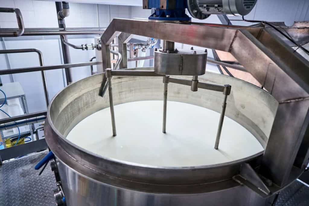 A large stainless steel industrial mixer with rotating blades stirs a vat filled with white liquid, likely milk, in a clean, modern dairy processing facility. Metal pipes and machinery surround the mixing tank.